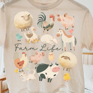 May include: Beige t-shirt featuring a colorful farm animal graphic. The design includes a sheep, rooster, pig, duck, horse, and cow, with the words "Farm Life!" printed below. The shirt is hanging on a wooden hanger.
