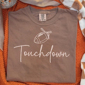 May include: A brown t-shirt with a white football graphic and the word "Touchdown" in script. The shirt is displayed on an orange blanket, with a disco ball and a checkered blanket in the background.