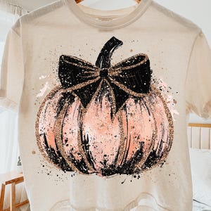 May include: Cream-colored t-shirt featuring a pink pumpkin design with a black bow and stem. The pumpkin has gold glitter accents and black splatter details, perfect for fall or Halloween.
