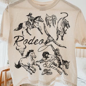 May include: Cream-colored t-shirt featuring a black and white rodeo-themed graphic. The design includes illustrations of cowboys, horses, bulls, and the words "Rodeo" and "8 Seconds". The shirt is hanging on a wooden hanger.