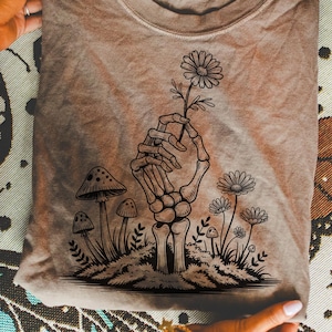 Op de afbeelding: Beige t-shirt met een zwarte illustratie van een skelet hand die een madeliefje vasthoudt. Het ontwerp bevat paddenstoelen en andere bloemen. Het shirt is opgevouwen en wordt door handen vastgehouden.