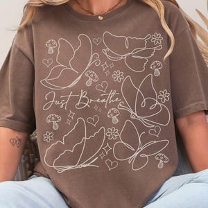 Puede incluir: Camiseta marr&oacute;n con un dise&ntilde;o de mariposas y flores blancas, y el texto "Just Breathe". El dise&ntilde;o incluye mariposas, corazones, flores y setas. La camiseta es de corte holgado.