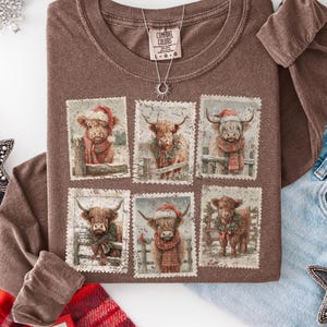May include: A brown long-sleeved top with a graphic of six Highland cows in festive Christmas outfits, presented as postage stamps. A silver necklace and a star-shaped decoration are also visible.