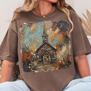 May include: Brown t-shirt featuring an artistic painting of a church with a steeple. The artwork has a textured, abstract style with blue, orange, and white accents. The shirt is a relaxed fit and has short sleeves.