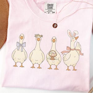 May include: A pale pink Comfort Colors t-shirt with a cartoon illustration of four geese. The geese are decorated with a bow, flower crown, basket of eggs, and bunny ears. A delicate necklace with a pendant is also visible.