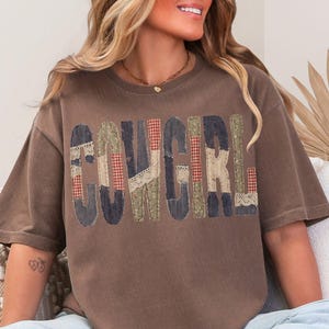 May include: Brown t-shirt with the word "COWGIRL" in patchwork letters. The letters are made of various patterned fabrics, including red plaid, blue denim, and cream lace. The shirt has short sleeves and a relaxed fit.