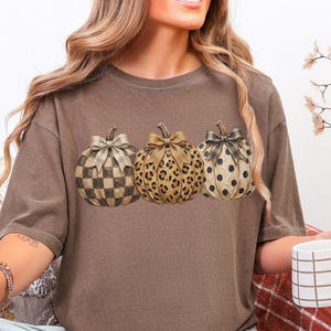 May include: A brown short-sleeve t-shirt featuring a graphic of three pumpkins arranged in a row. Each pumpkin is decorated with a different pattern, including a checkered print, a leopard print, and a polka dot print, each topped with a bow.