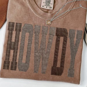 May include: A brown Comfort Colors t-shirt with the word "HOWDY" in patchwork letters. The shirt has a crew neck and a small tag. A silver necklace with a pendant is draped across the shirt. A brown hat is visible in the upper left corner.