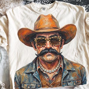 May include: A cream-colored t-shirt featuring a graphic illustration of a person wearing a brown cowboy hat, leopard print sunglasses, a thick silver-toned chain necklace, and a denim jacket with a prominent dark mustache on their face.