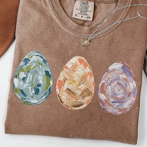 Peut inclure: T-shirt marron avec trois &oelig;ufs de P&acirc;ques peints en bleu, orange et violet. Les &oelig;ufs pr&eacute;sentent un motif textur&eacute; au pinceau. Un collier argent&eacute; avec un pendentif est pos&eacute; sur le t-shirt.
