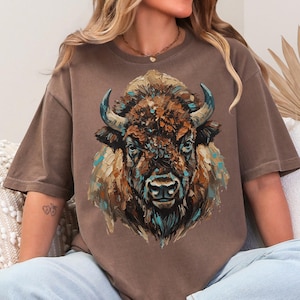 May include: Brown t-shirt featuring a detailed bison illustration. The artwork uses a palette of browns, blues, and oranges, creating a textured, painterly effect. The shirt has short sleeves and a relaxed fit, suitable for casual wear.