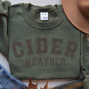 K&ouml;nnte beinhalten: Ein olivgr&uuml;nes Sweatshirt mit den Worten "CIDER WEATHER" in braunen Blockbuchstaben aufgedruckt. Ein brauner Hut, Jeans und ein karierter Schal sind ebenfalls im Bild.