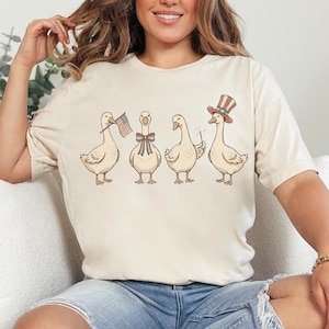 May include: Beige t-shirt featuring a whimsical illustration of four geese. Each goose is adorned with patriotic accessories: a small American flag, a bow tie, a sparkler, and a top hat. The design is printed on a soft, comfortable tee.
