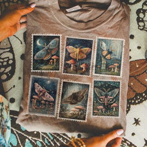 Puede incluir: Una camiseta color topo con seis ilustraciones estilo sello postal de polillas y hongos. La obra de arte utiliza azules, verdes y amarillos sobre un fondo oscuro y estrellado. La camiseta está doblada, mostrando las ilustraciones detalladas.