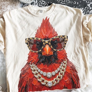 May include: Cream-colored t-shirt featuring a detailed illustration of a red cardinal bird wearing leopard print sunglasses and a gold chain necklace with diamond accents. The shirt has a crew neck.
