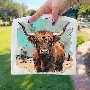 May include: White t-shirt featuring a detailed illustration of a Highland cow with long horns and shaggy brown fur. The design includes a desert landscape with cacti and a turquoise and gray background.