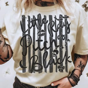 May include: A cream-colored t-shirt featuring a black graphic print of vertical stripes and the text "Little Ray of Pitch Black." The design includes small illustrations of a pair of lips, a spider, a skull, a bow, and bats in black ink.