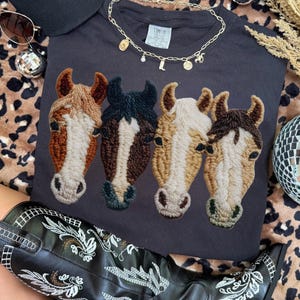 May include: Dark gray t-shirt featuring embroidered horse heads in shades of brown, black, and cream. A gold necklace with charms, a black hat with white text, sunglasses, and a pair of black cowboy boots with white embroidery are also visible.