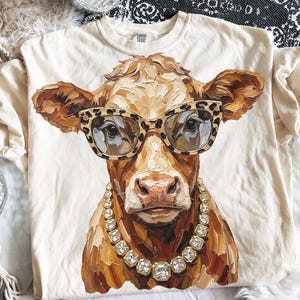 以下が含まれることがあります： クリーム色の半袖Tシャツ。中央には、ヒョウ柄のメガネと大ぶりのラインストーンのネックレスを身につけた茶色の牛のグラフィックが描かれています。牛のイラストは、筆致が見える質感のある絵画風のスタイルで表現されています。