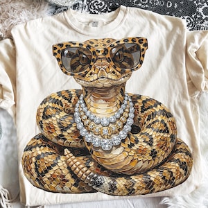 Puede incluir: Camiseta color crema con una ilustración detallada de una serpiente con gafas de sol con estampado de leopardo y un collar de diamantes. La serpiente está enrollada, con un patrón de escamas marrones, beige y negras. La camiseta es de material suave.
