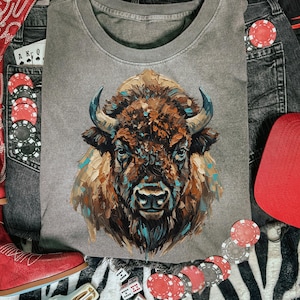 May include: Gray crew neck t-shirt featuring a detailed bison illustration in shades of brown, blue, and tan. The shirt is surrounded by a red cap, playing cards, poker chips, dice, and red cowboy boots, creating a western theme.