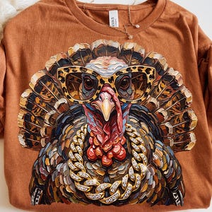 May include: A rust-colored t-shirt featuring a graphic print of a turkey wearing leopard-print glasses and a thick, sparkling gold chain. The turkey has detailed, textured feathers in shades of brown, black, and tan, with a bright red wattle.
