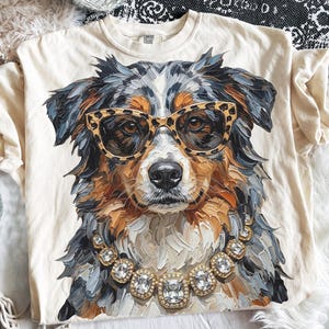 Pode incluir: Uma t-shirt cor de creme com o gráfico de um cão pastor australiano a usar óculos com padrão de leopardo e um colar de strass grosso. A ilustração utiliza pinceladas espessas e texturizadas em tons de cinzento, preto e castanho claro.