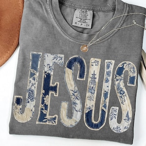 May include: Gray Comfort Colors t-shirt with the word "JESUS" appliquéd in blue and cream floral fabric. A gold necklace with a pendant rests on the shirt. A brown hat is visible in the upper left corner.