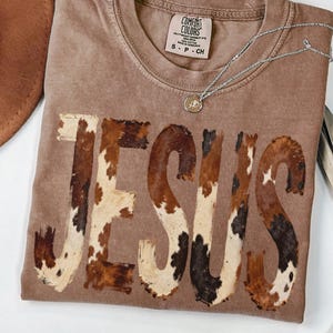 May include: A brown Comfort Colors t-shirt with the word "JESUS" in a cowhide pattern. A silver necklace with a pendant rests on the shirt. A brown hat is partially visible in the upper left corner.