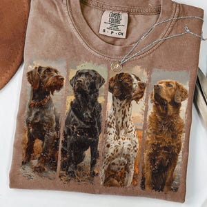 May include: A brown Comfort Colors t-shirt featuring a graphic of four different dog breeds. A silver necklace with a pendant rests on the shirt. The shirt is folded on a white surface, with a brown hat in the upper left corner.