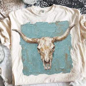 May include: Cream-coloured t-shirt with a detailed illustration of a cow skull with long horns. The skull is painted in shades of white, brown, and beige, set against a textured teal background. The shirt is a Comfort Colors brand.