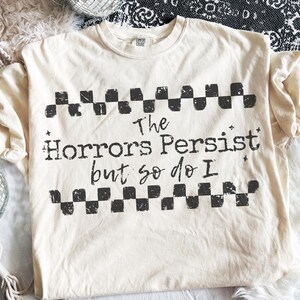 May include: Cream-colored t-shirt with the text "The Horrors Persist but so do I" in black, framed by a checkered pattern. The shirt is a Comfort Colors brand.