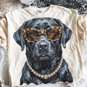 May include: A cream-colored t-shirt featuring a graphic of a black Labrador retriever wearing leopard print sunglasses and a gold-toned rhinestone chain necklace. The dog illustration has visible brushstroke textures and a detailed, realistic appearance.