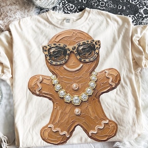May include: A cream-colored t-shirt featuring a central graphic of a brown gingerbread person. The gingerbread figure wears leopard print sunglasses and a gold-toned chain necklace with hexagonal links. The design has a textured, painted appearance.
