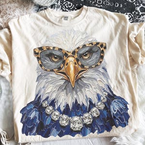 May include: A cream-colored t-shirt featuring a graphic of a bald eagle wearing leopard print glasses and a large, multi-faceted clear crystal necklace. The eagle has white head feathers and dark blue chest feathers, rendered in a textured, painterly style.