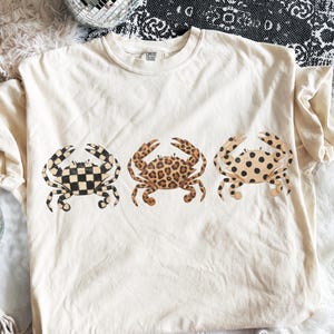 以下が含まれることがあります： 3匹のカニのデザインが施されたクリーム色のTシャツ。カニは、白黒のチェッカーボード、ヒョウ柄、黒い斑点のあるタン色のカニというユニークなパターンです。このシャツはComfort Colorsブランドです。