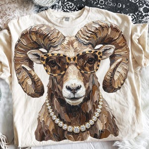 May include: A cream-colored t-shirt featuring a central graphic of a ram with large, textured, brown curled horns. The ram is wearing leopard-print sunglasses and a necklace made of round, clear, faceted stones, rendered in an impasto paint style.