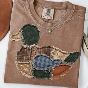 May include: A brown short-sleeved t-shirt featuring a large duck-shaped appliqué made from various fabric patches including plaid, floral, and corduroy patterns with frayed edges. The inner neck label reads COMFORT COLORS, 100% COTTON, MADE IN HONDURAS, S P CH.