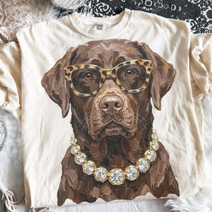 May include: A cream-colored t-shirt featuring a painterly illustration of a brown Labrador retriever wearing leopard print glasses and a chunky rhinestone necklace. The dog is depicted with visible brushstrokes in shades of brown and tan.