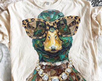 May include: A cream-colored t-shirt featuring a graphic of a mallard duck wearing leopard print glasses and a gold chain with clear square stones. The duck is rendered in a thick, textured impasto paint style with shades of green, brown, and yellow.