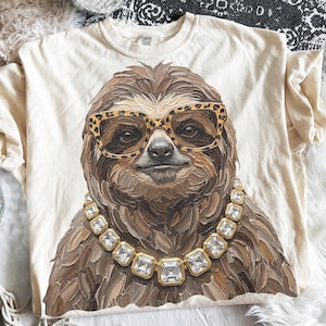 Puede incluir: Una camiseta de color crema con un gráfico central de un perezoso marrón con detalles de pinceladas texturizadas. El perezoso lleva gafas con estampado de leopardo y un collar de cadena dorada con grandes cristales cuadrados transparentes.