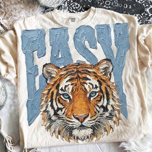 May include: Cream-colored t-shirt with the word "EASY" in light blue, textured lettering. The shirt features a detailed tiger face with orange, black, and white fur, and blue eyes. The shirt is a Comfort Colors brand.