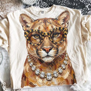 May include: Cream-colored t-shirt featuring a cougar wearing leopard print glasses and a diamond necklace. The cougar's portrait is painted in shades of brown, tan, and gold. The shirt is made of a soft material.