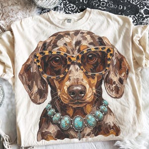 Peut inclure: Un t-shirt couleur crème arborant une illustration graphique d'un teckel tacheté marron portant des lunettes à motif léopard et un collier de pierres turquoise. Le chien présente un aspect texturé avec des coups de pinceau visibles.