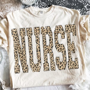 May include: Cream-colored t-shirt with the word "NURSE" in large, bold letters. The letters are filled with a leopard print pattern. The shirt has short sleeves and a crew neck.