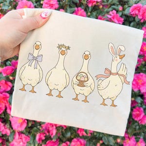 May include: A cream tote bag with four cartoon geese. One has a lavender bow, another a floral crown, a third carries a basket of Easter eggs, and the last wears bunny ears and a pink bow. The background is a blur of pink flowers.