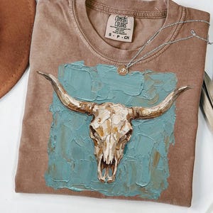May include: A brown Comfort Colors t-shirt featuring a bull skull design on a teal background. The bull skull is painted in shades of gold and cream. A silver necklace with a small pendant is draped across the shirt.