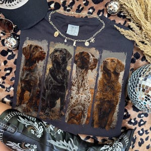 May include: A black t-shirt featuring four different dog breeds in a vertical rectangular design. The shirt is accessorized with a gold necklace and a black and white baseball cap. A pair of sunglasses and a disco ball are also visible.
