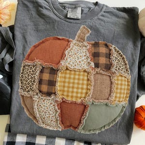 May include: A charcoal gray crew neck t-shirt featuring a large, central pumpkin graphic made of various fabric patches. The pumpkin design includes squares of brown plaid, leopard print, yellow gingham, floral patterns, and solid orange and sage green.