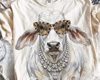 May include: A cream-coloured t-shirt featuring a graphic illustration of a cow wearing leopard print sunglasses and a multi-layered crystal necklace. The cow is rendered in a textured, painterly style with visible brushstrokes in shades of white, grey, and tan.